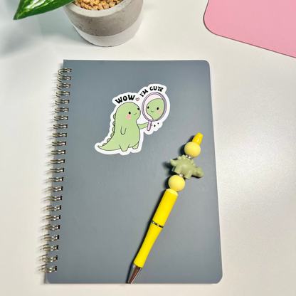 A gray spiral notebook with a cute green dinosaur sticker that says 'WOW, I'M CUTE' lies on a white desk next to a yellow pen with a dinosaur charm. A potted plant and a pink item are partially visible in the background.