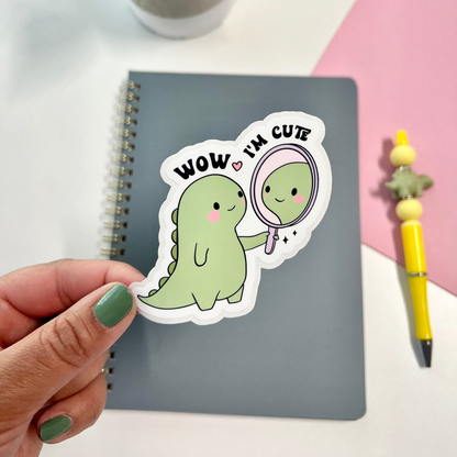 A hand with green nail polish holds a die-cut sticker featuring a cute light green dinosaur holding a mirror with the text 'WOW I'M CUTE' in black. The sticker is positioned over a gray spiral notebook on a white desk, with a pink background section, a yellow pen, and a potted plant visible.