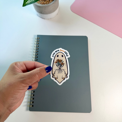 A hand with dark blue nail polish holds a die-cut sticker above a gray spiral notebook on a white desk. The sticker features a cute ghost illustration holding a bouquet of colorful flowers, with the text 'you are bootiful' in a white arch above it.