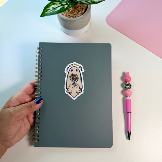 A hand holds a dark gray spiral notebook with a decorative sticker on the cover, placed on a white desk next to a pink pen and a potted plant.