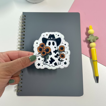 Notebook with a decorative sticker featuring a ghost and sunflowers, held by a hand on a pink and white background.